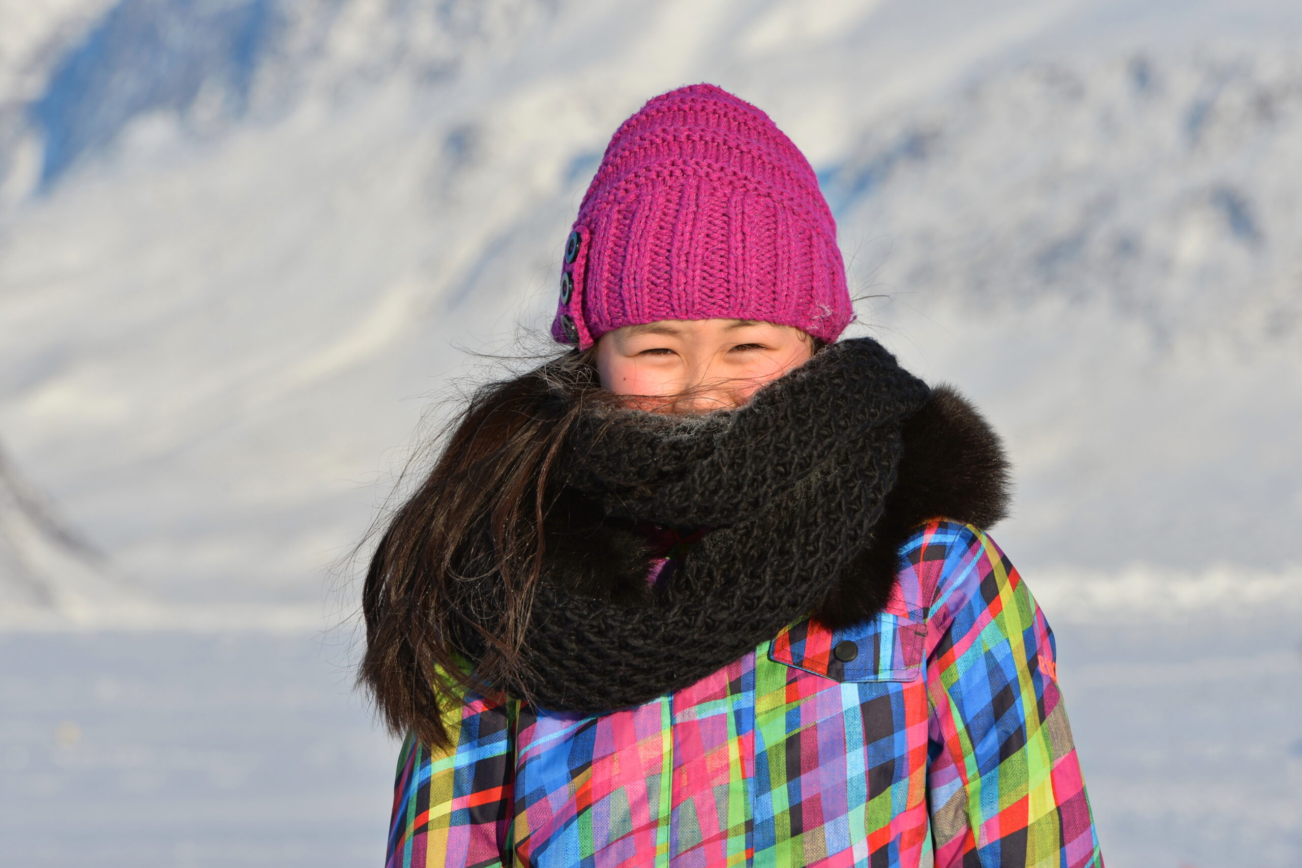 Photo of a young person wearing a pink knitted hat and a dark brown scarf which is covering most of their face. They are standing in front of a snowy background.